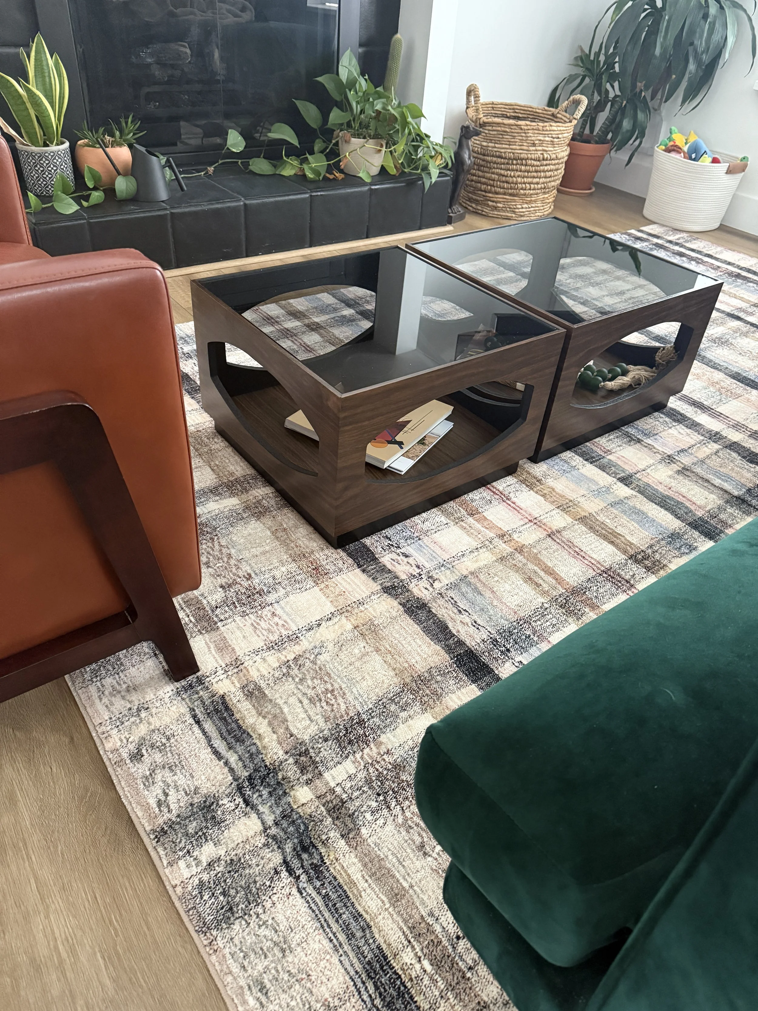 Living room with a black fireplace, potted plants, a glass coffee table, a patterned area rug, an orange armchair, a green velvet seat, and a white basket filled with toys.