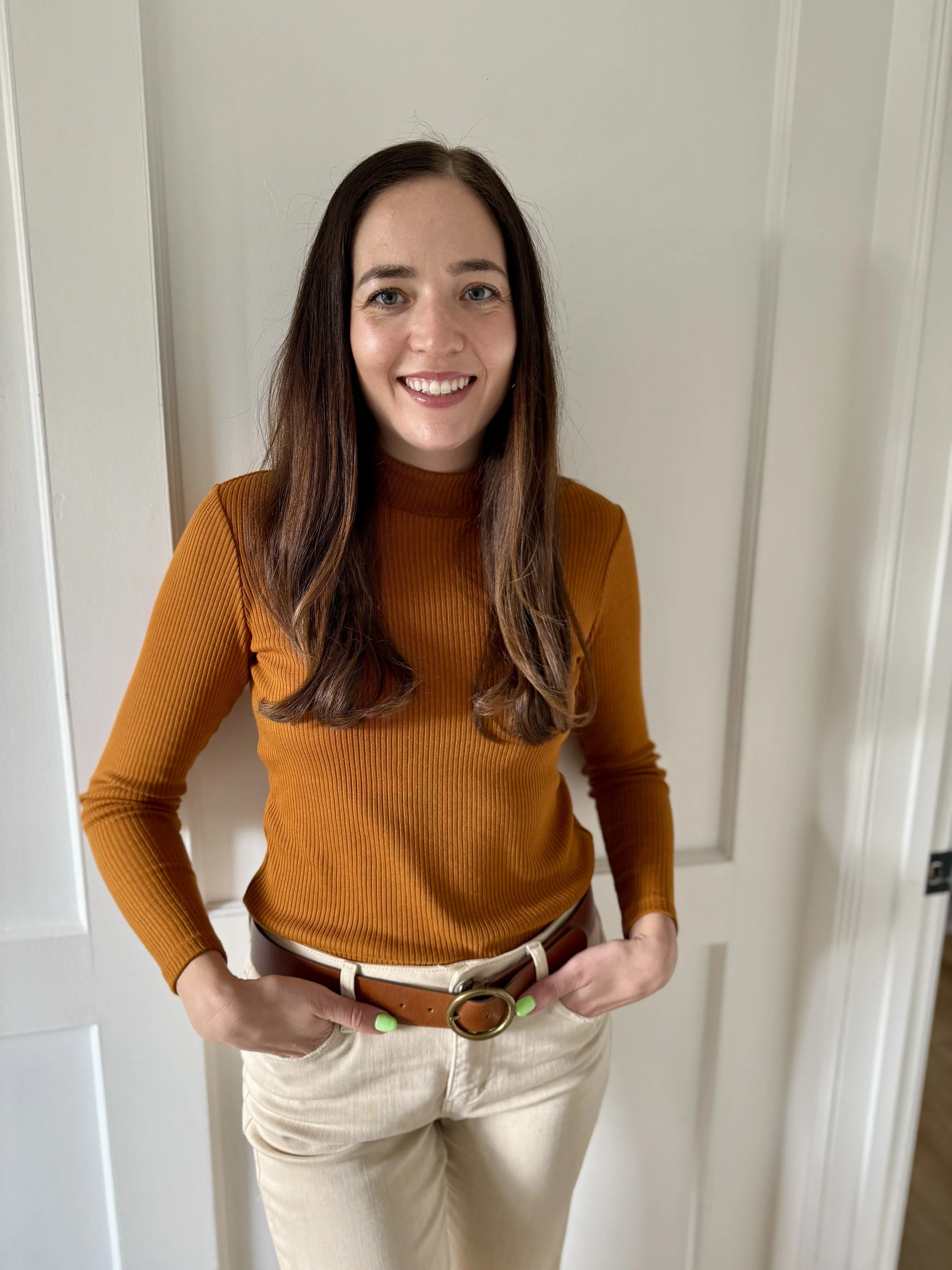 A woman with long brown hair, blue eyes, and light skin is smiling; she's wearing a rust-colored ribbed turtleneck sweater, beige pants, and a brown belt with a gold buckle, standing against a plain off-white wall.