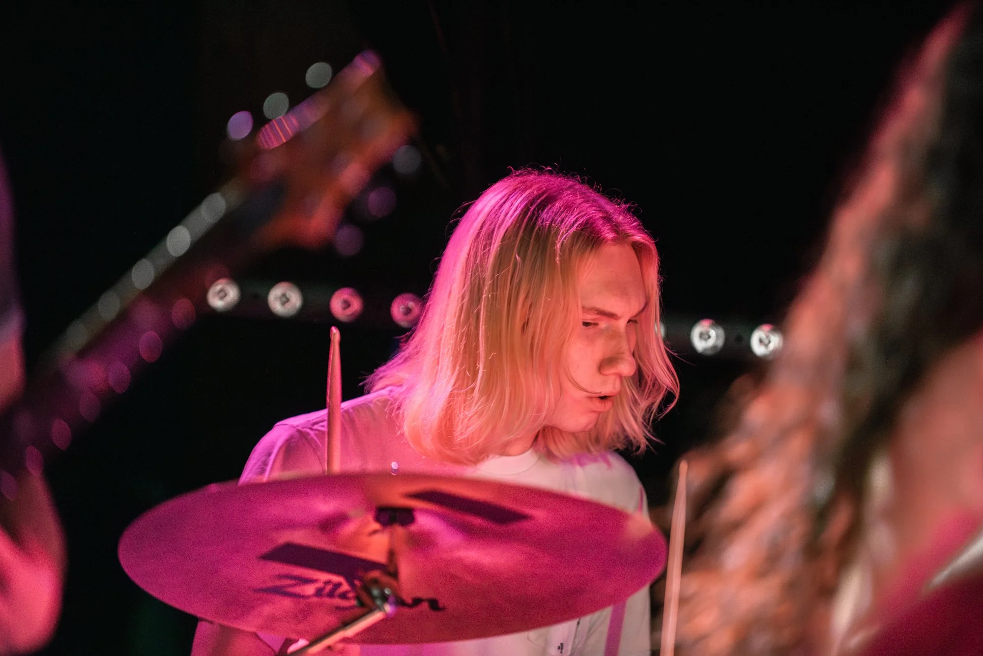 A young man with long blonde hair playing drums during a live music performance, illuminated by pink stage lighting.
