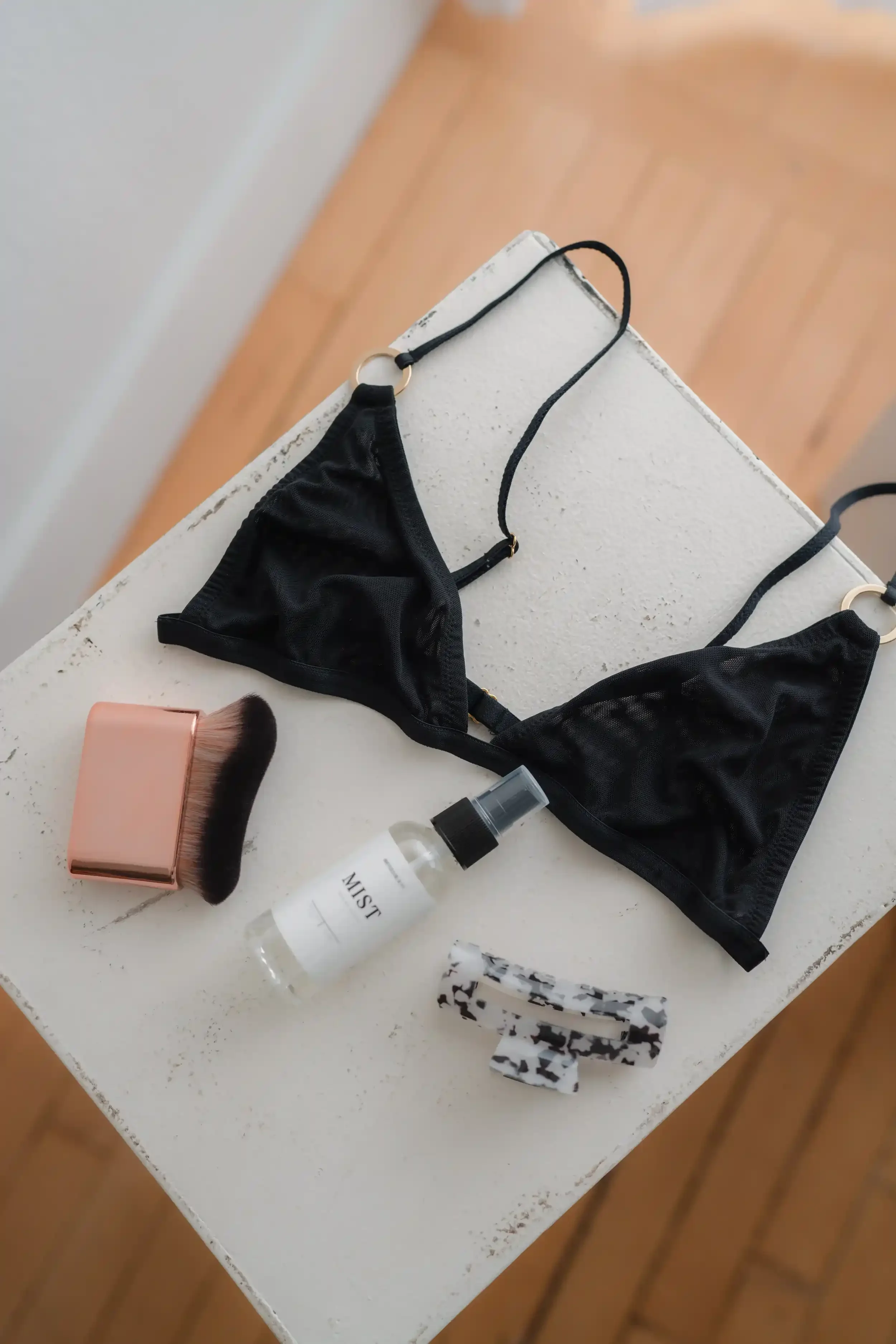 A black string bikini top on a white surface, a small spray bottle labeled 'MIST', a makeup brush with a pink handle, a black and white patterned hair clip, and a black and white patterned scrunchie.