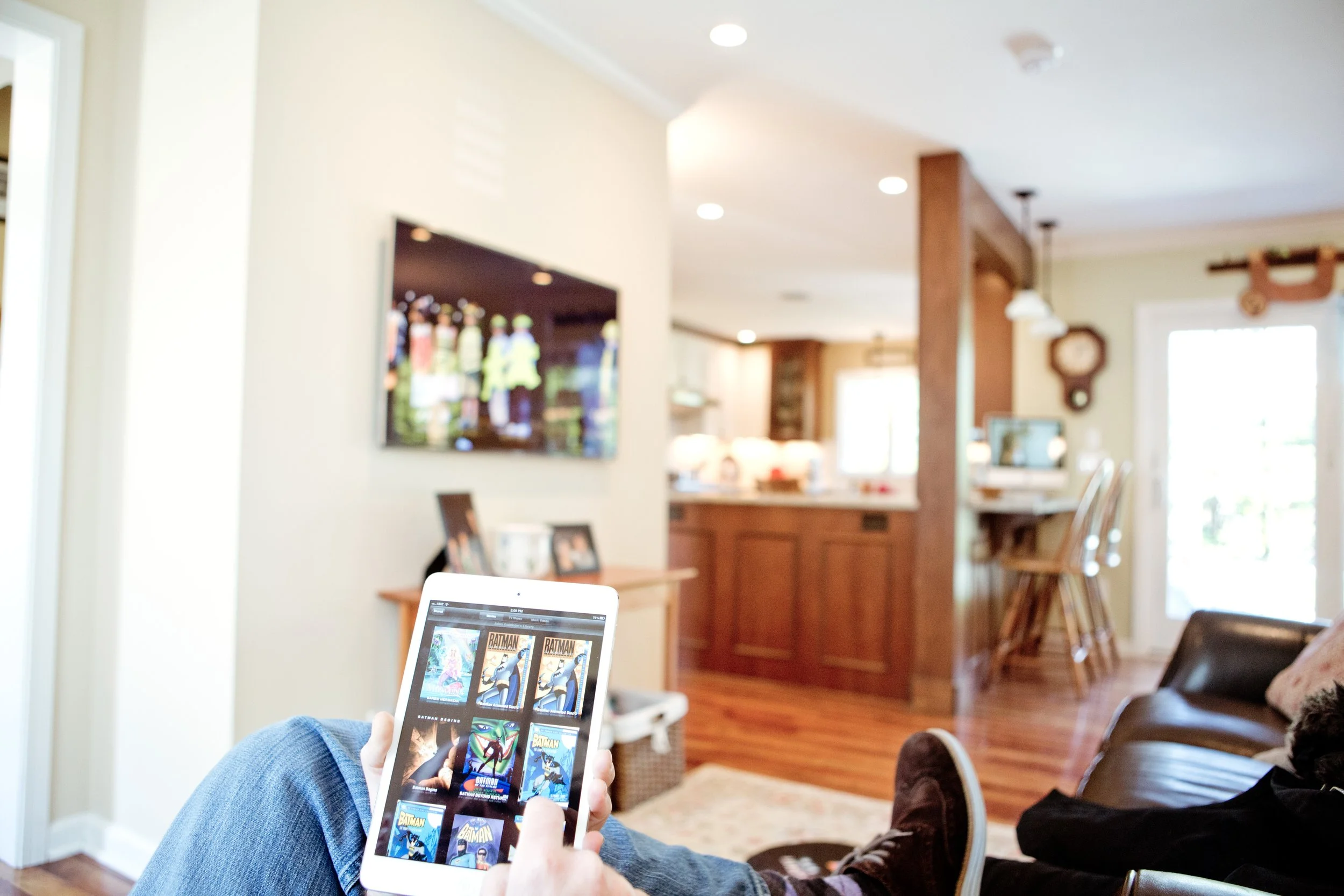 Person sitting on a couch in a living room, using a tablet to browse comic books, with a TV on the wall and a kitchen in the background.