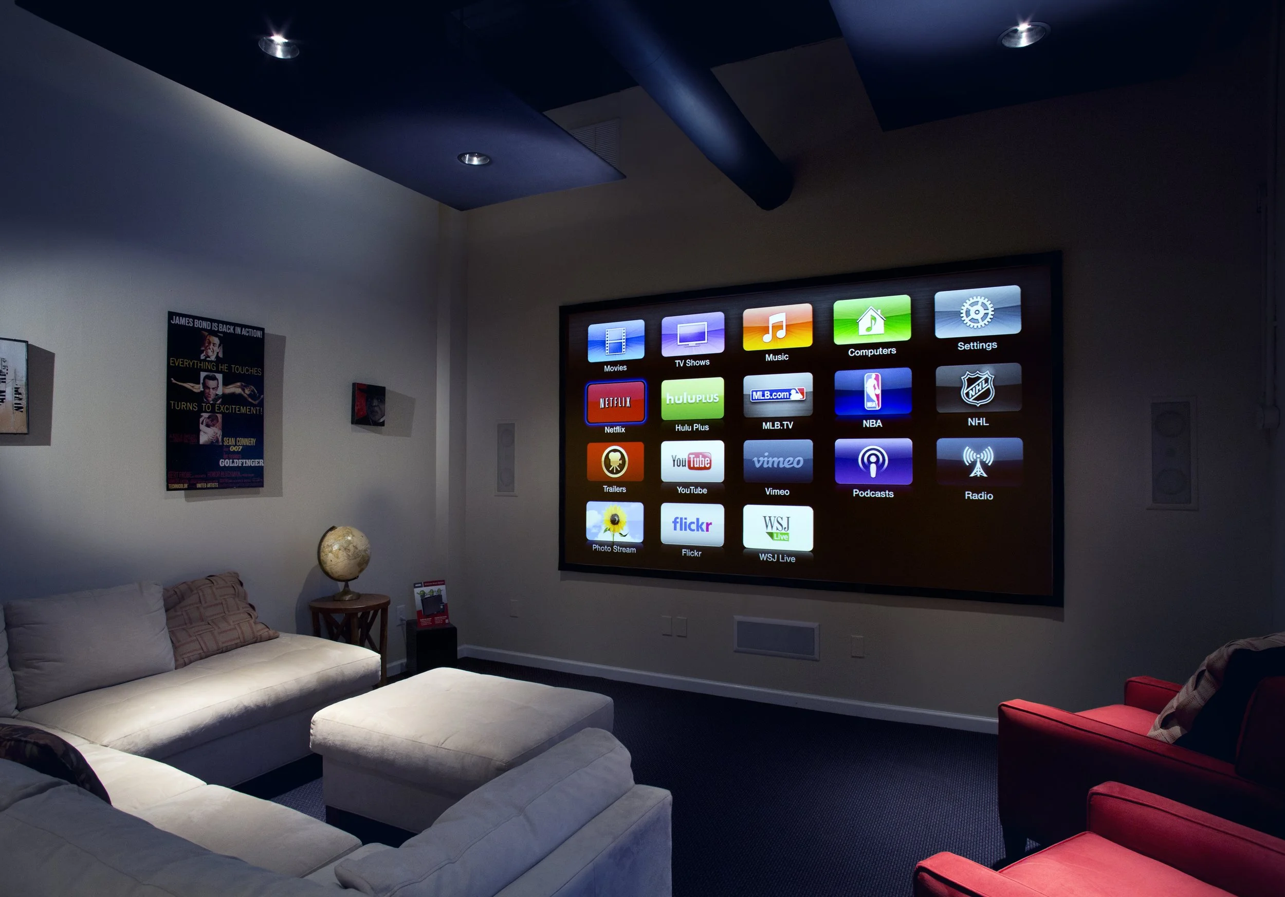 Home theater room with a large screen showing streaming app icons, white couch, red armchair, and wall posters.