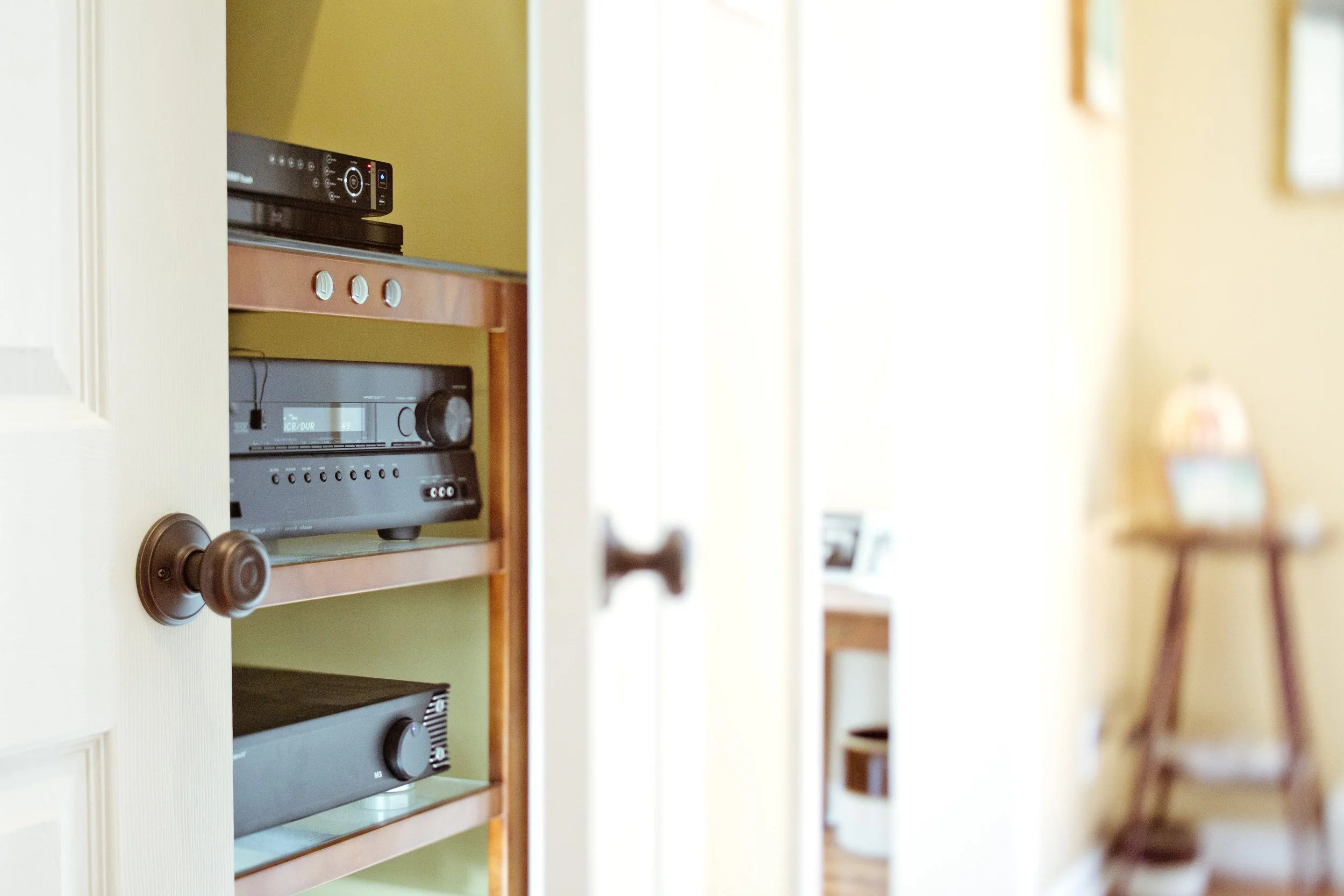 Partially open door revealing a wooden shelf with audio equipment inside a room with yellow walls and a small table with a lamp and picture frames in the background.