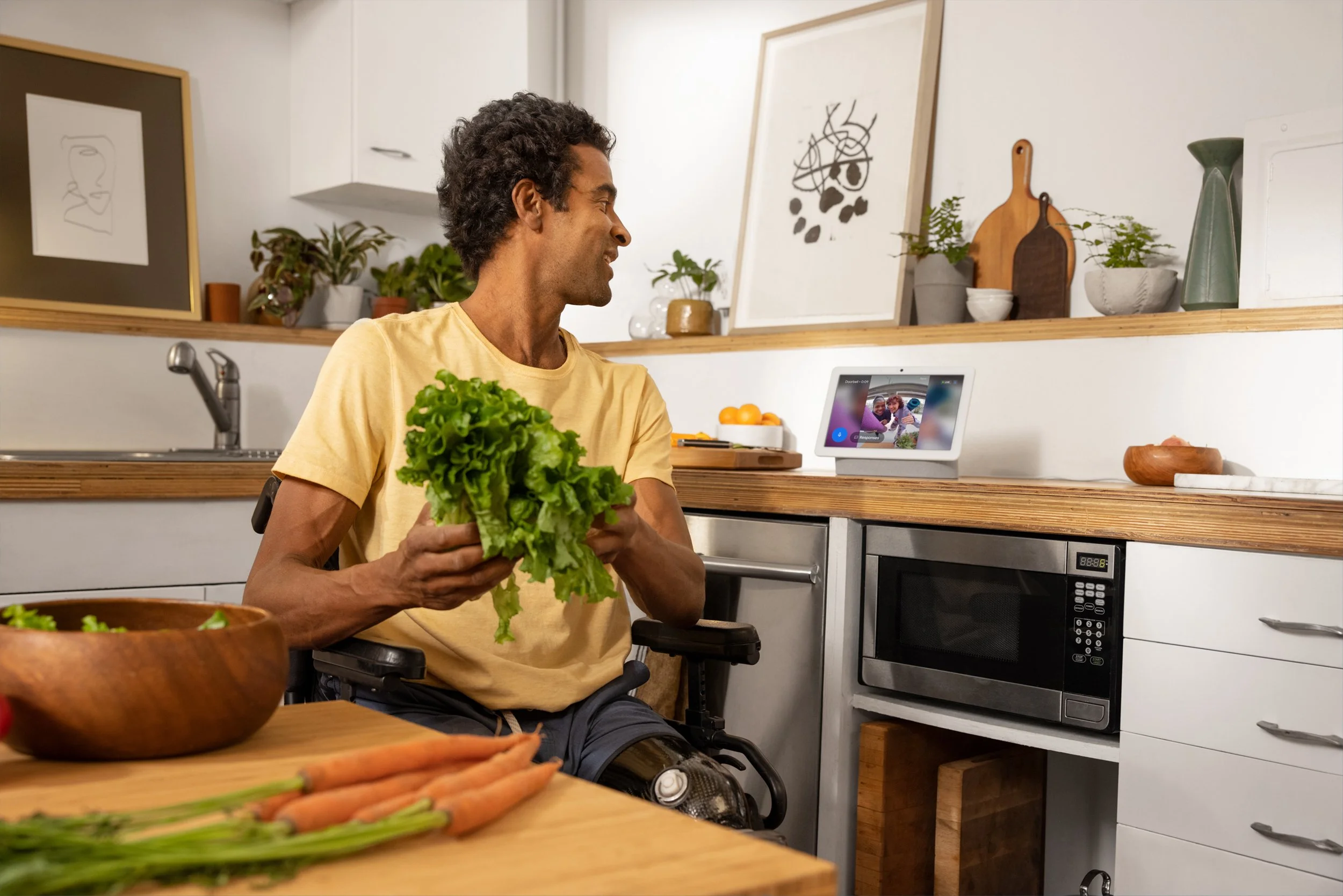 A man in a wheelchair holding a head of lettuce in a kitchen with a wooden countertop and modern appliances, looking at a smart home device on the counter.