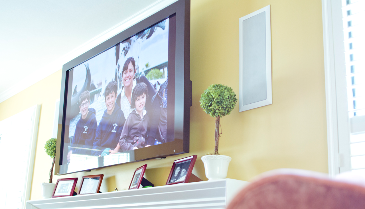 A living room with a large mounted flat-screen TV showing a woman and three boys outdoors. The wall is painted yellow and decorated with framed photographs, a potted topiary plant, and a rectangular wall panel. Natural light streams in from a window with white blinds on the right.