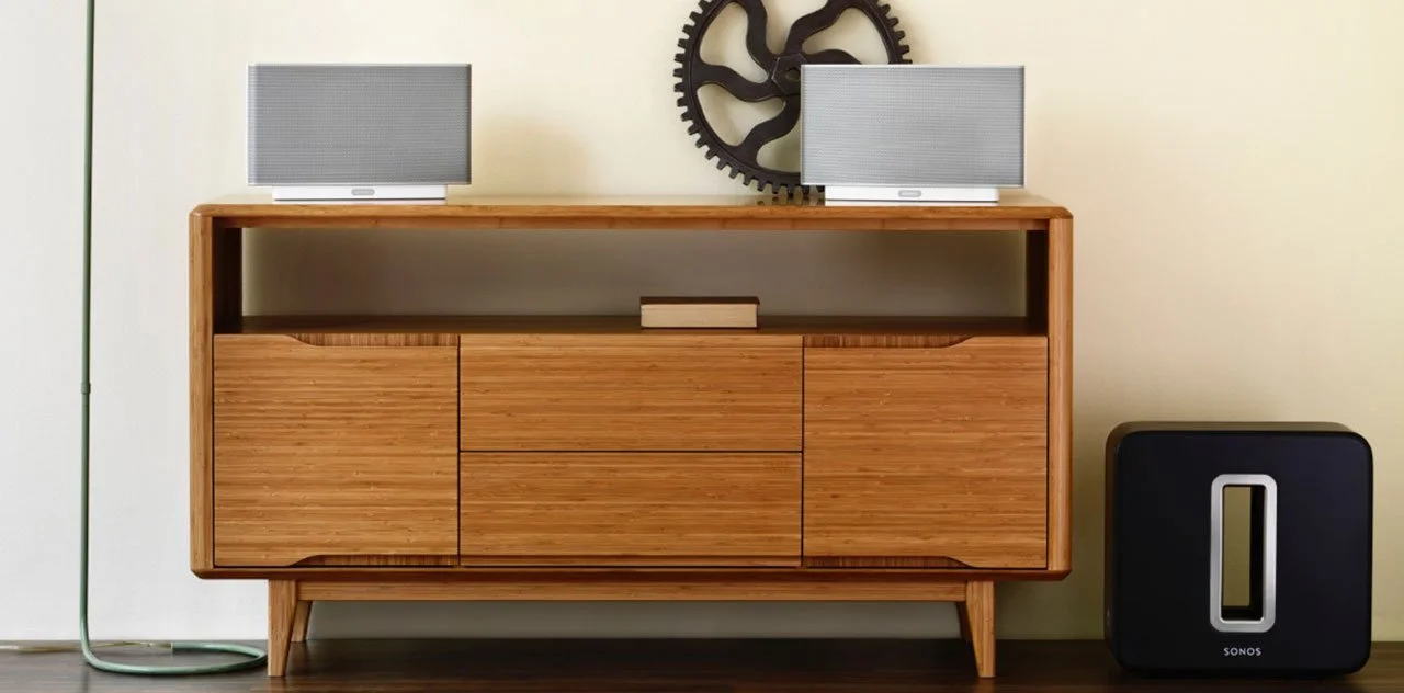 A wooden sideboard with two speakers on top, a decorative gear wall art, a small book, a black subwoofer, and a tall green floor lamp to the left.