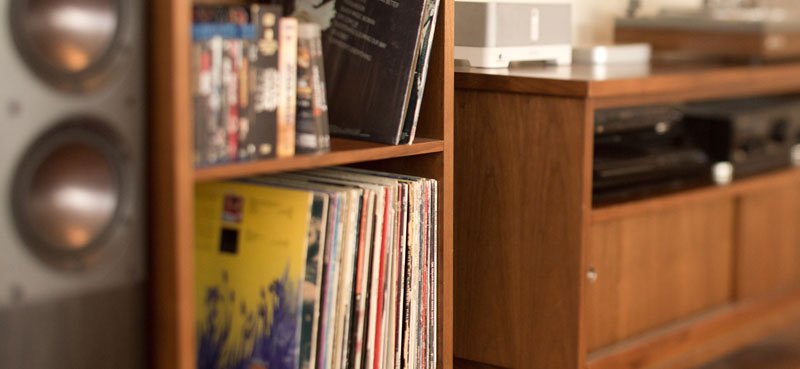 A wooden bookshelf filled with vinyl records, a black speaker in the background, and a wooden TV stand with electronic devices.