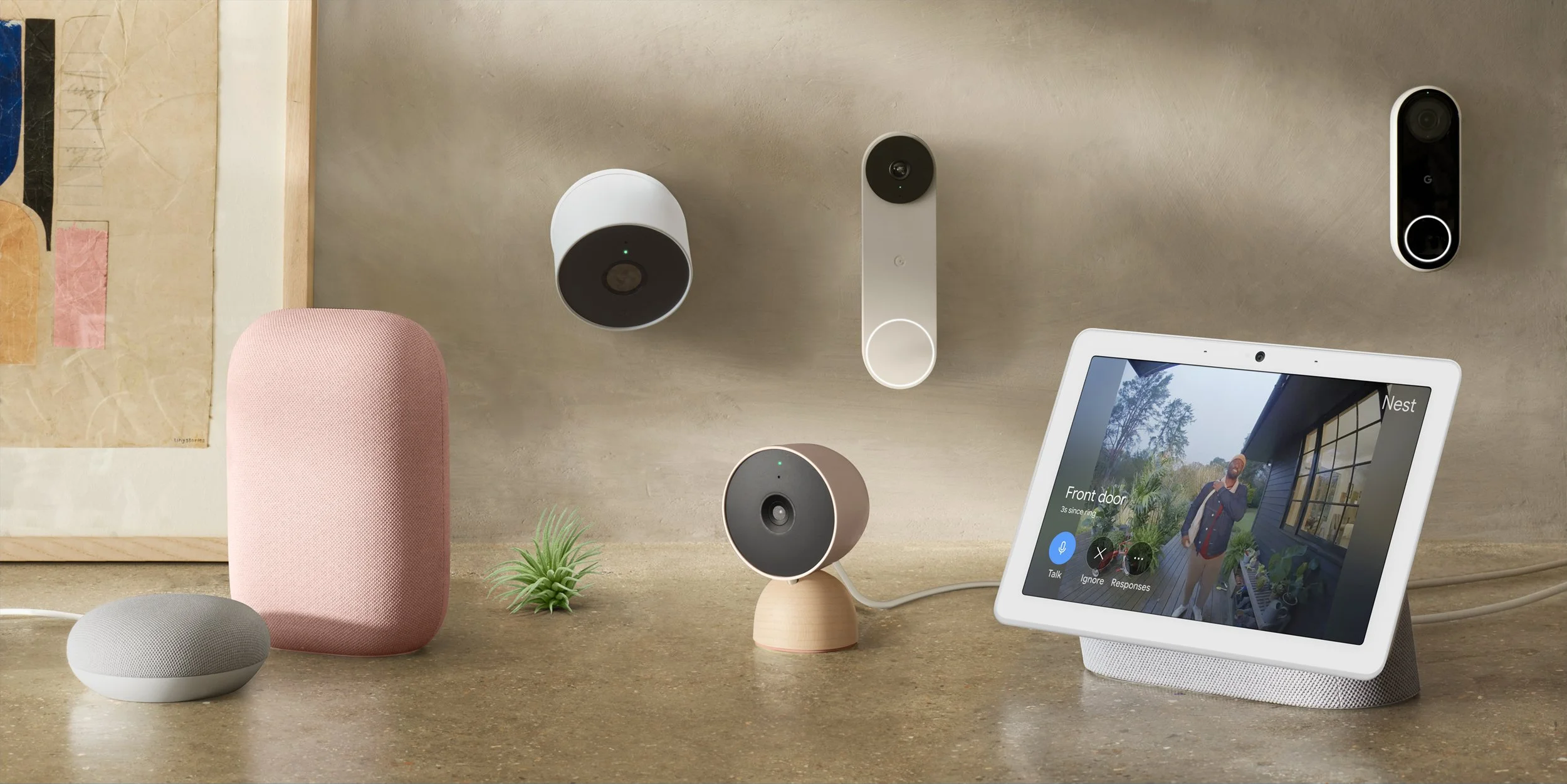 Home security system setup with smart cameras, a smart display showing a front door view, a pink smart speaker, a small gray smart speaker, a tiny potted plant, and a piece of artwork in the background.