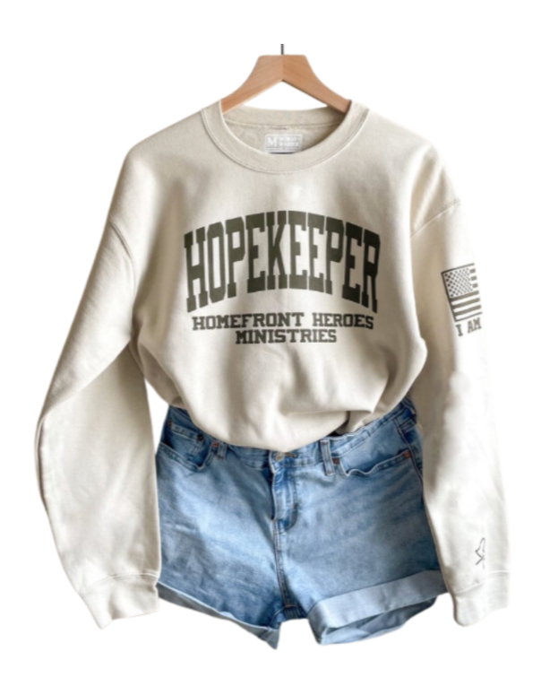 Cream-colored sweatshirt with the word 'HOPEKEEPER' and 'HOMEFRONT HEROES MINISTRIES' printed on it, hanging on a wooden hanger with denim shorts below.