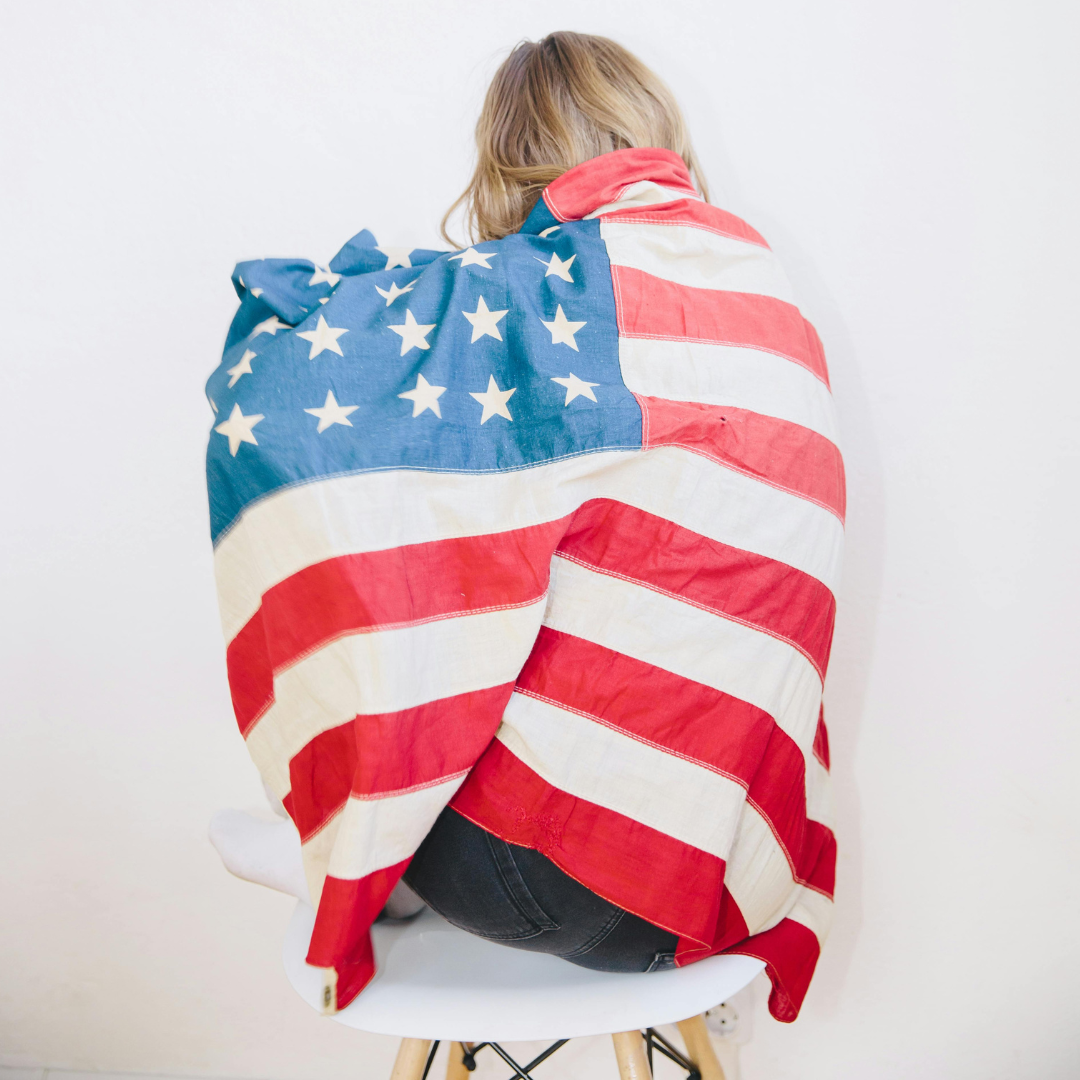Person sitting on a white stool, seen from back, wrapped in a large American flag.