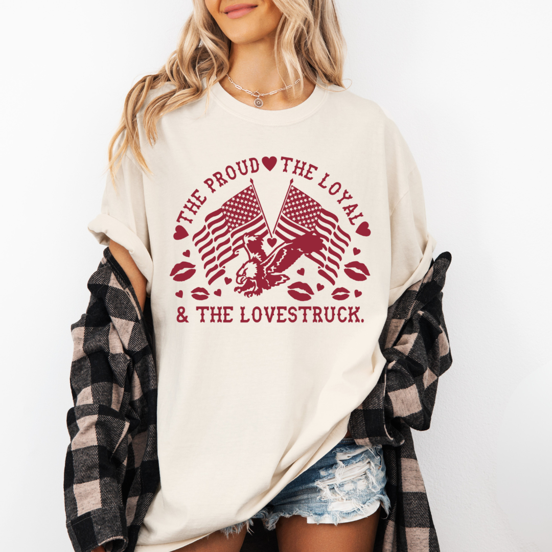 A woman wearing a white t-shirt with red printed graphics of American flags, an eagle, and the words 'The Proud, The Loyal & The Lovestruck', along with red hearts and lips, paired with a black and beige checkered shirt tied around her waist and distressed denim shorts.
