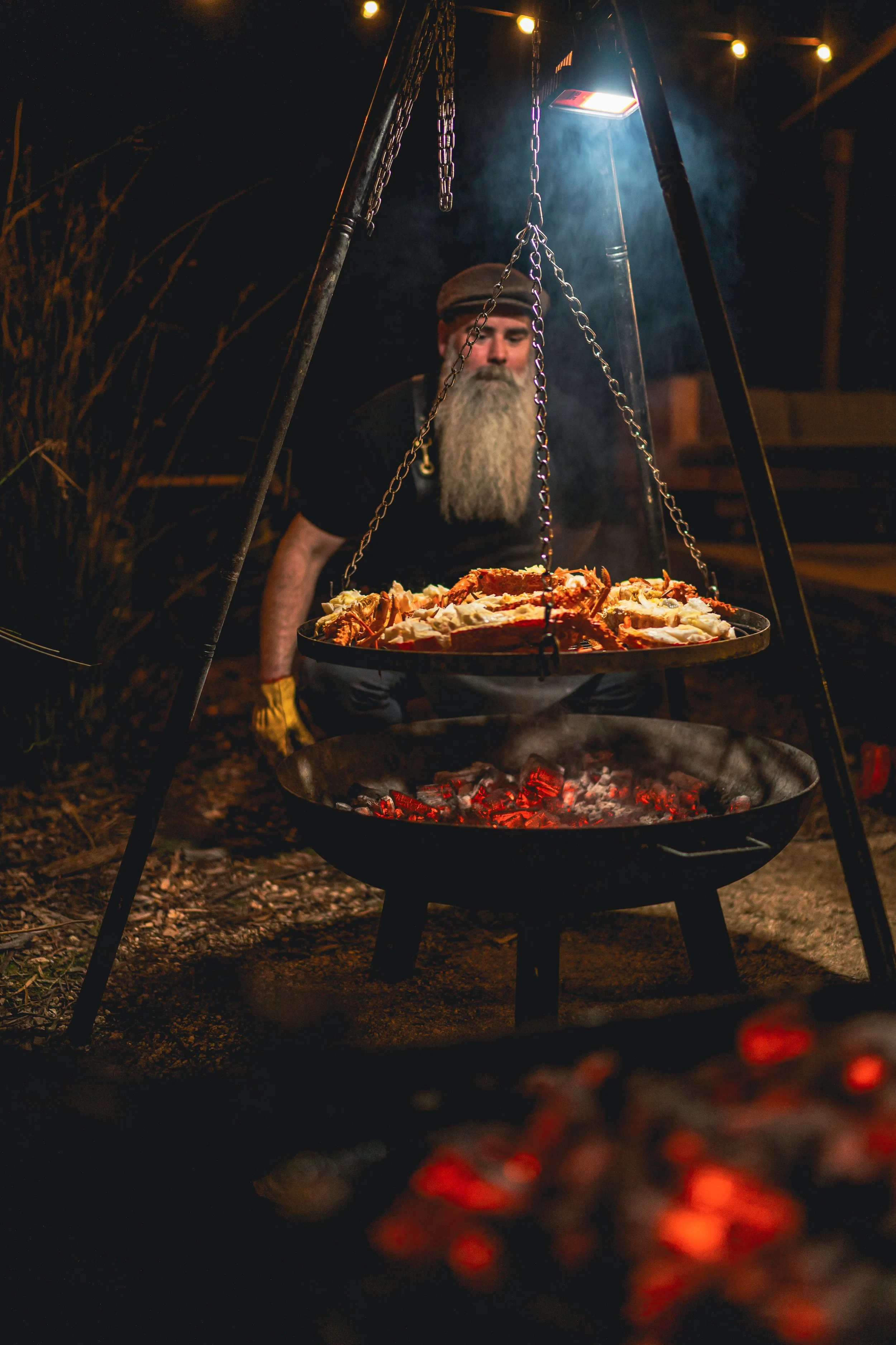 A man with a long gray beard wearing a hat, preparing to serve food from a hanging tray over a campfire grill at night.