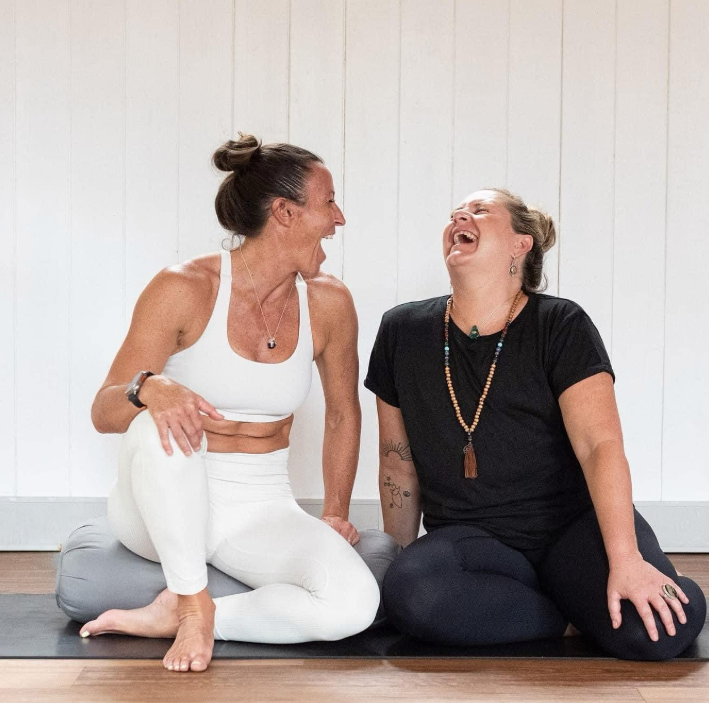 Two women are sitting on yoga mats against a white wall, laughing and enjoying each other's company.