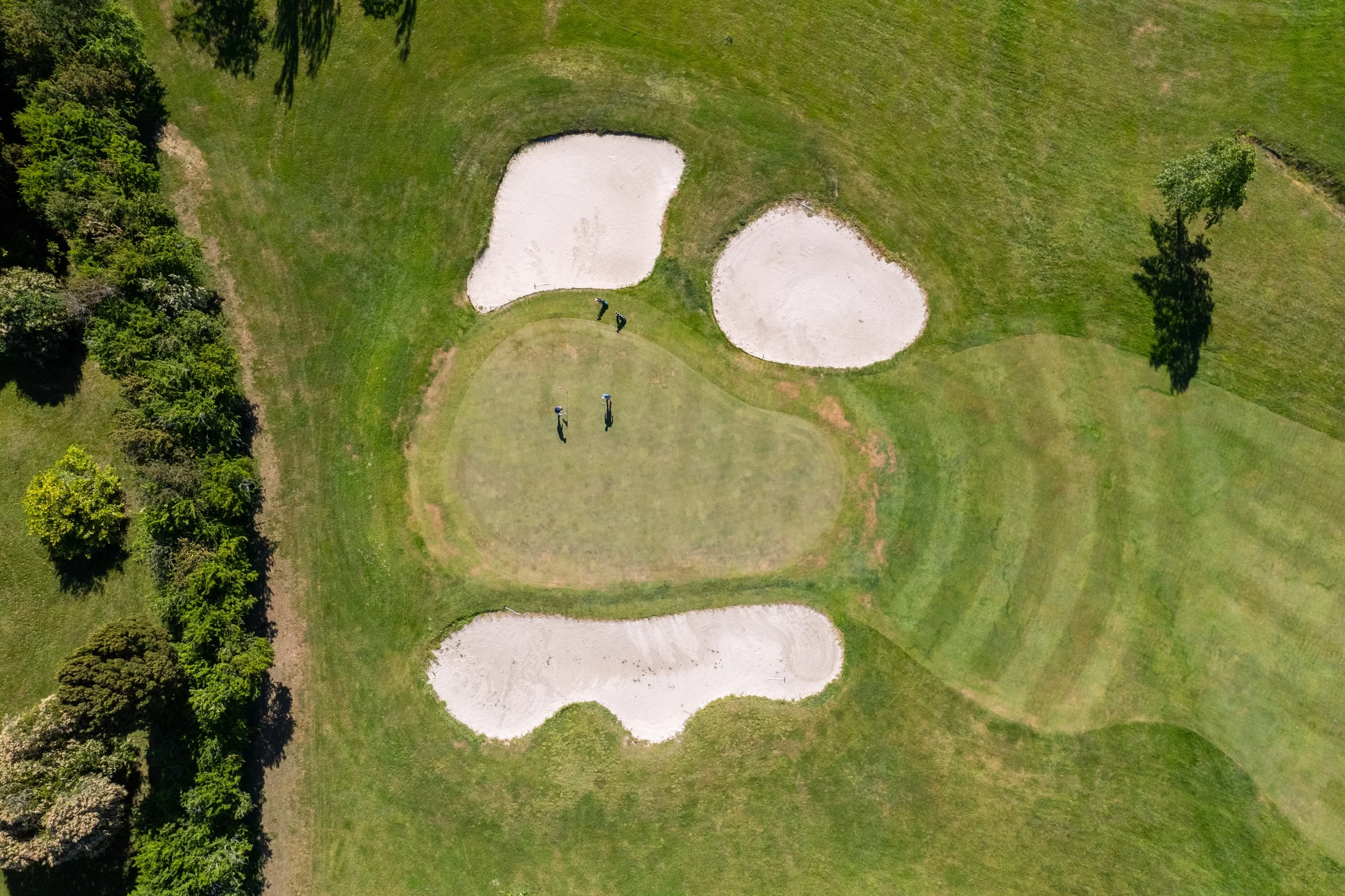 Aerial view of a golf course with two sand bunkers and three people on the green, surrounded by grass and trees.