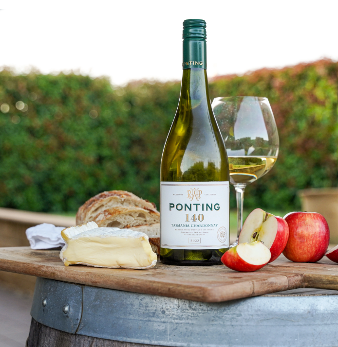 A bottle of Ponting 140 Tasmanian Chardonnay wine, a glass of white wine, a wedge of brie cheese, a loaf of bread, and sliced apples on a wooden surface outdoors.