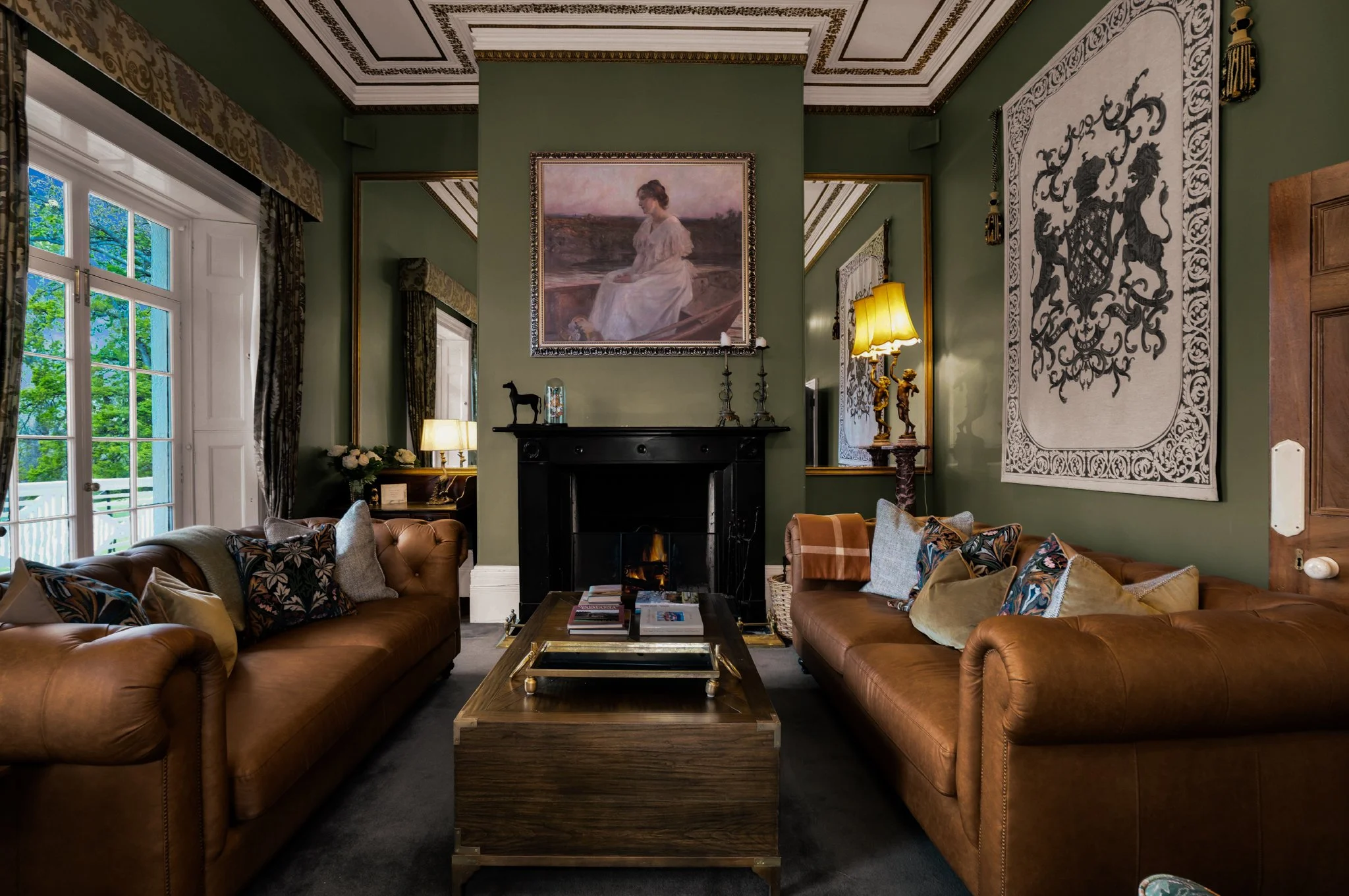 Living room with green walls, gold-trimmed ceiling, and large windows with patterned curtains. There are two brown leather sofas, a wooden coffee table with books, and a black fireplace with decorative items. Several framed artworks and a tapestry ar