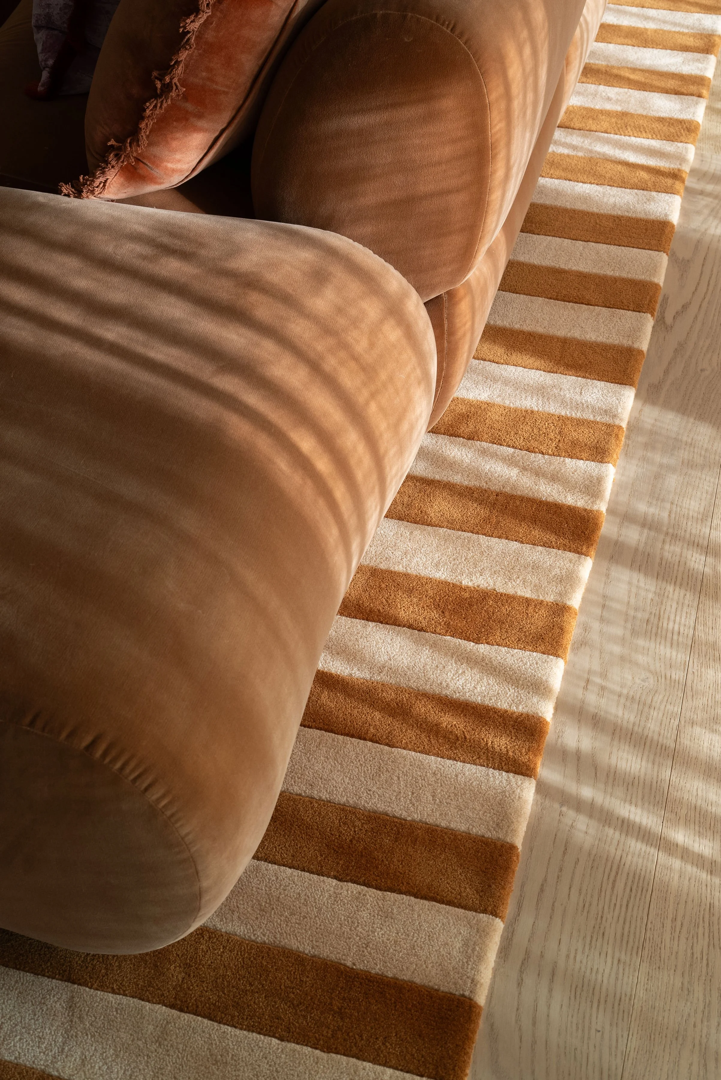 A brown leather sofa with rounded armrest, positioned on a rectangular rug with beige and brown stripes, on a light wooden floor.