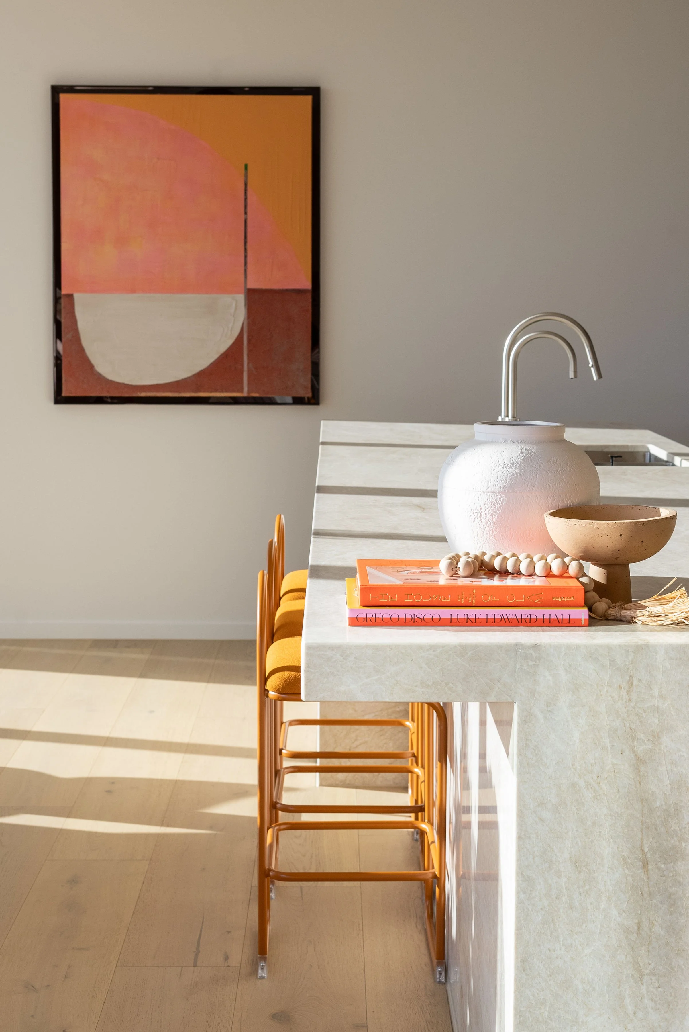 A modern kitchen with a white marble island, orange chairs, and abstract wall art. Decor includes books, a ceramic bowl, a beaded necklace, and a large white textured vase with a faucet.