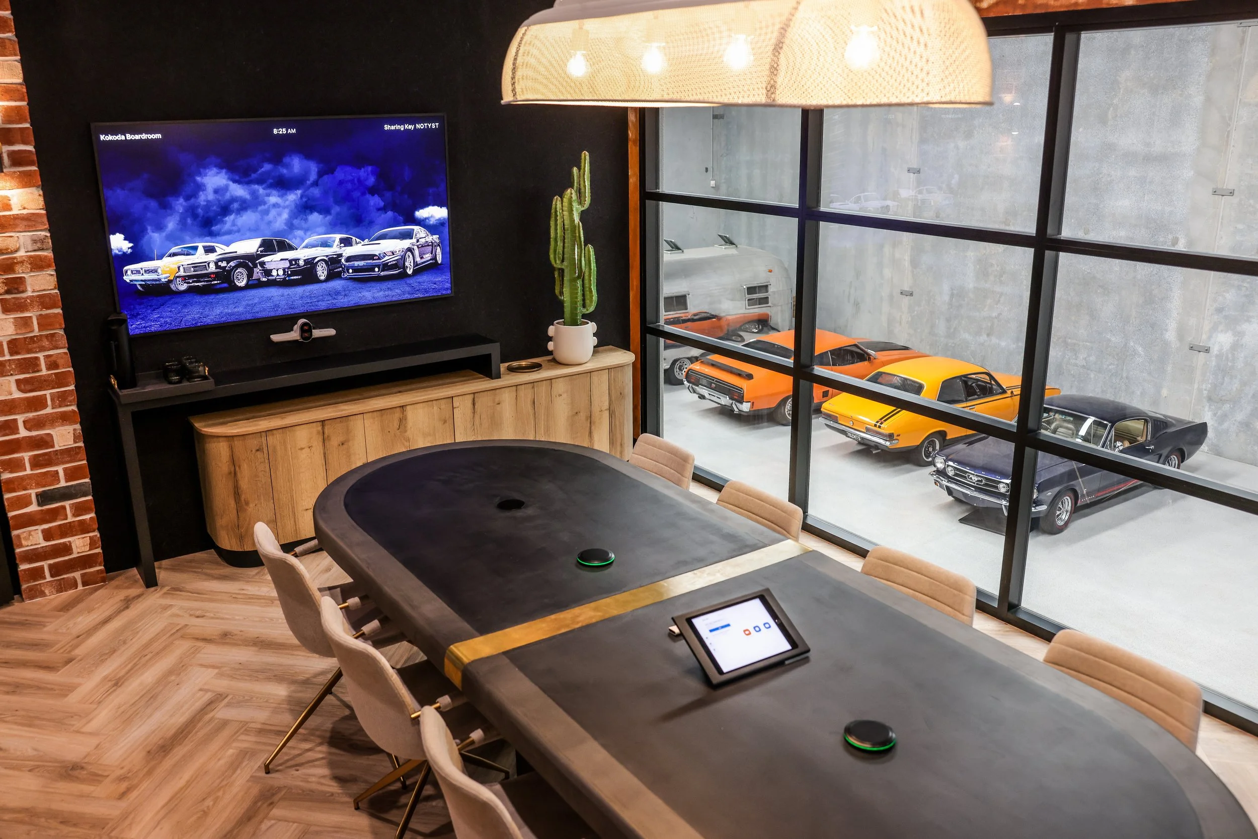 Modern conference room with a large dark table, beige chairs, and a wall-mounted TV displaying classic cars. Large windows reveal a garage with vintage muscle cars in yellow, black, and orange, and a silver trailer.