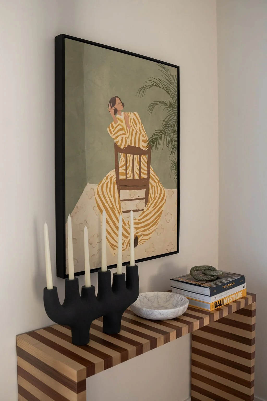 A framed illustration of a woman with dark hair wearing a yellow and white striped dress, sitting on a chair with one hand resting on her face and the other behind her head, in front of a green wall with a plant, hanging on a wall in a room with a wooden striped console table decorated with a black candle holder with five white candles, a marble bowl, and books.