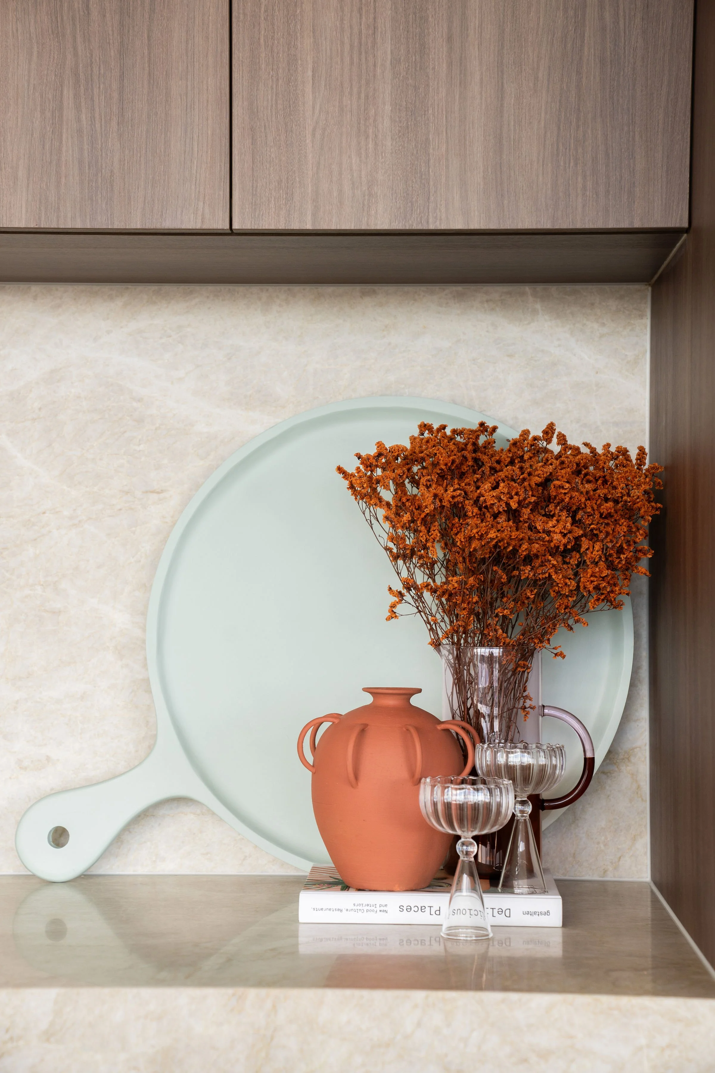 Decorative kitchen countertop display with a large mint-green cutting board, an orange terracotta vase with dried orange flowers, two clear glass goblets, and a book.