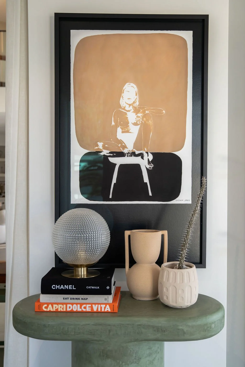A framed abstract artwork featuring a woman sitting on a stool, with a nude color background, hung on a dark wall. Below the artwork is a green table with a textured globe lamp on a stack of black and white books, a beige ceramic vase, a small beige pitcher, and a potted plant with long, spiny leaves.