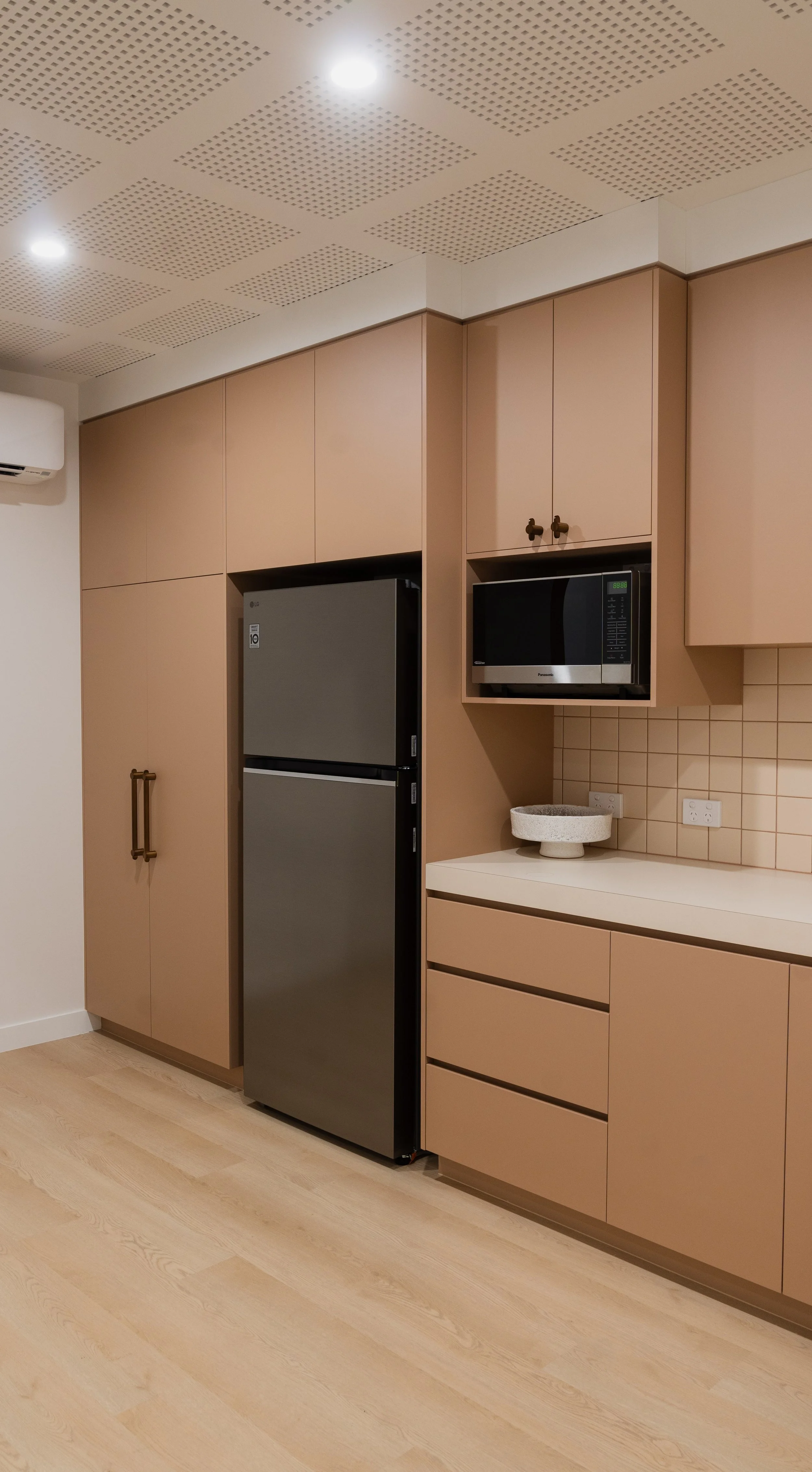 Modern kitchen with beige cabinets, a gray refrigerator, a microwave, and a countertop with a decorative bowl, on light wooden flooring.