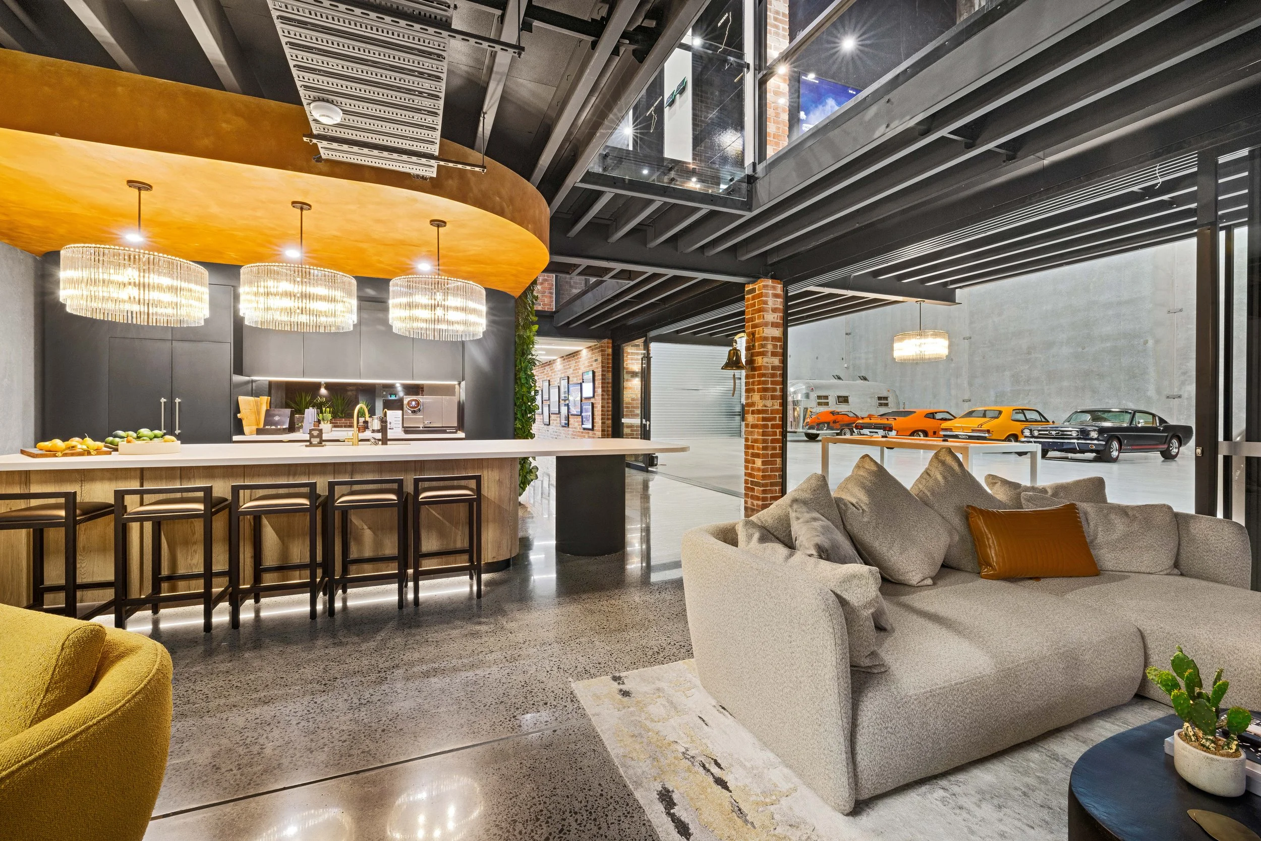 Modern indoor lounge with a bar area, beige sofa with pillows, and vintage cars visible through large glass windows.