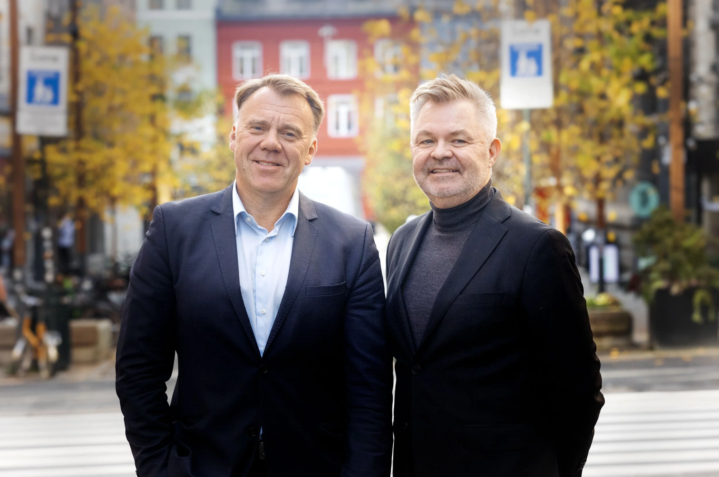 Two men in suits standing outdoors on a city street during autumn, smiling at the camera.