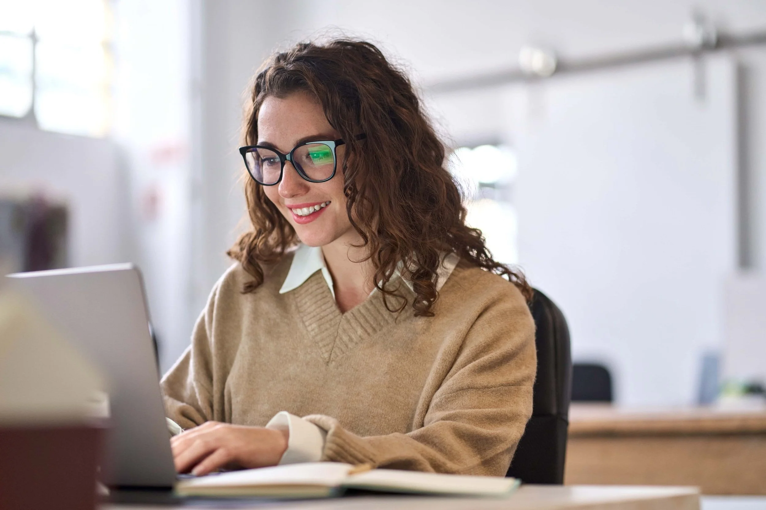 A woman with curly brown hair, glasses, and a beige sweater working on a laptop in an office.