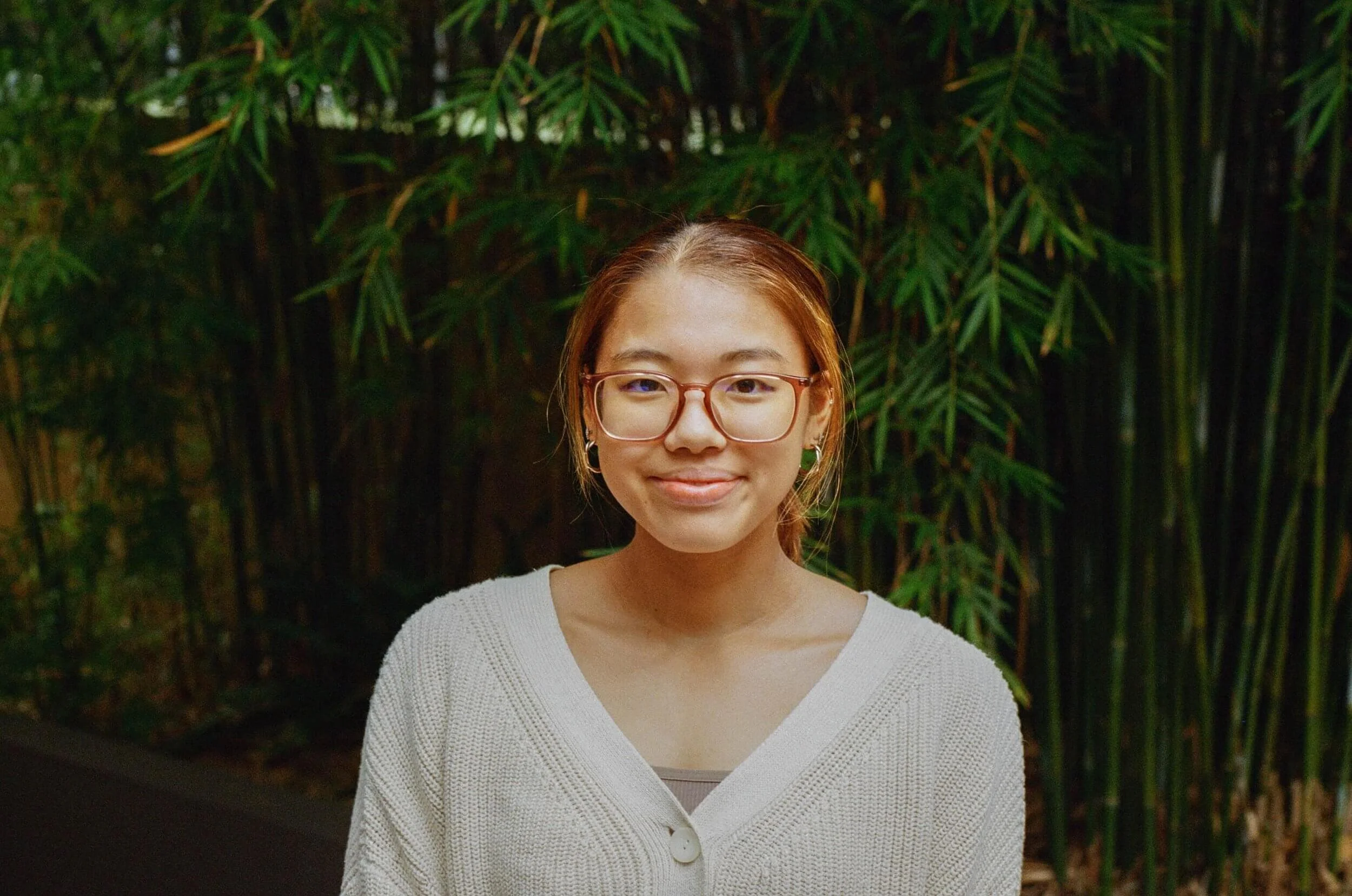 A young woman with brown hair, glasses, and hoop earrings standing in front of greenery, wearing a white cardigan and smiling softly.