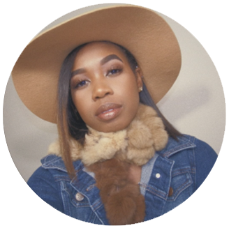 Photo of Re- brown woman with a brown hat, scarf, and jean jacket on looks neutral in her facial expression in front of a grey background.