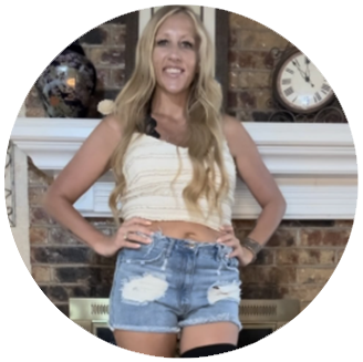 Photo of Angie- white woman with long blonde hair wearing a pale yellow tank crop top and jean shorts stands in front of a fireplace smiling with her hands on her hips.