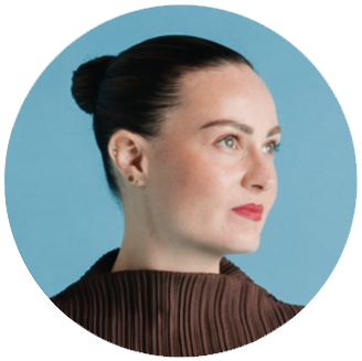Photo of Molly Burke- white woman smiles with her mouth closed. Her hair is dark and tied up in a bun. She is wearing a brown shirt and looking to the right in front of a blue background.