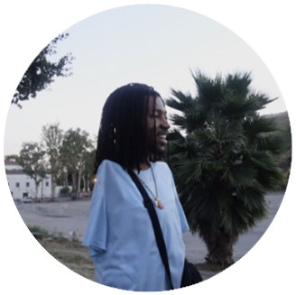 Photo of Mir- short black man with long dark hair wearing a white shirt smiles around trees outside.