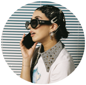 Photo of April Lockhart: white woman with hair up wearing clips in hair and a stylish beaded collared shirt, holding her phone to her ear in front of a black and white striped backdrop