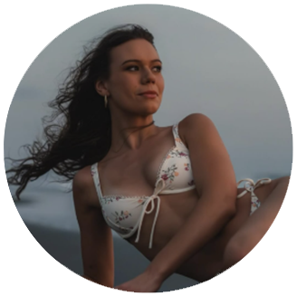 White woman with curly long dark hair and a white bikini with flowers on it lays on a cloudy dark beach.