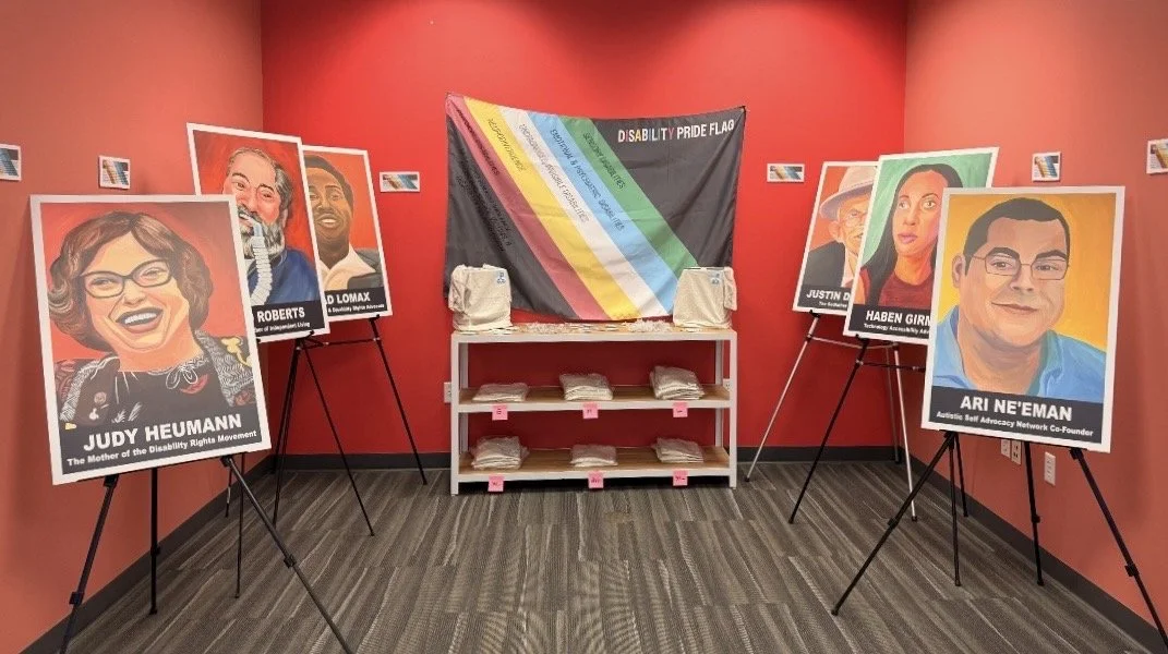 Photo of 6 disability activists on posters sitting on tripods surrounding a disability pride flag with free tshirts in front of it in front of a red wall.