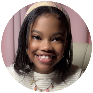 Photo of Ar’miah-black girl smiling with shoulder length hair and headband wearing white striped shirt and homemade necklace in front of a pink background