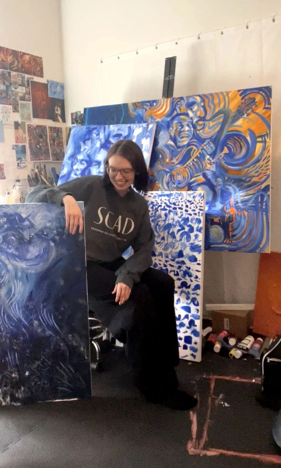 Emma in her studio sitting in front of 3 blue paintings, holding 1 blue painting.