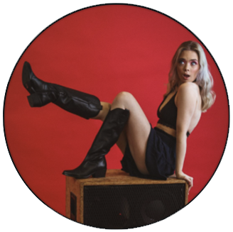 Photo of Kitty- White woman with long blonde hair wearing black tank crop top, shorts, and black boots, sits on top of a crate with 1 leg up in front of a bright red background.