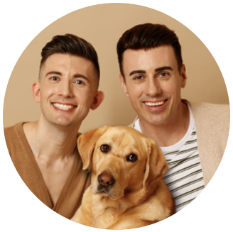 Photo of Matthew & Paul Castle & Guide Dog Maple- 2 white men with short brown hair smile with their golden retriever guide dog in front of a light brown backdrop