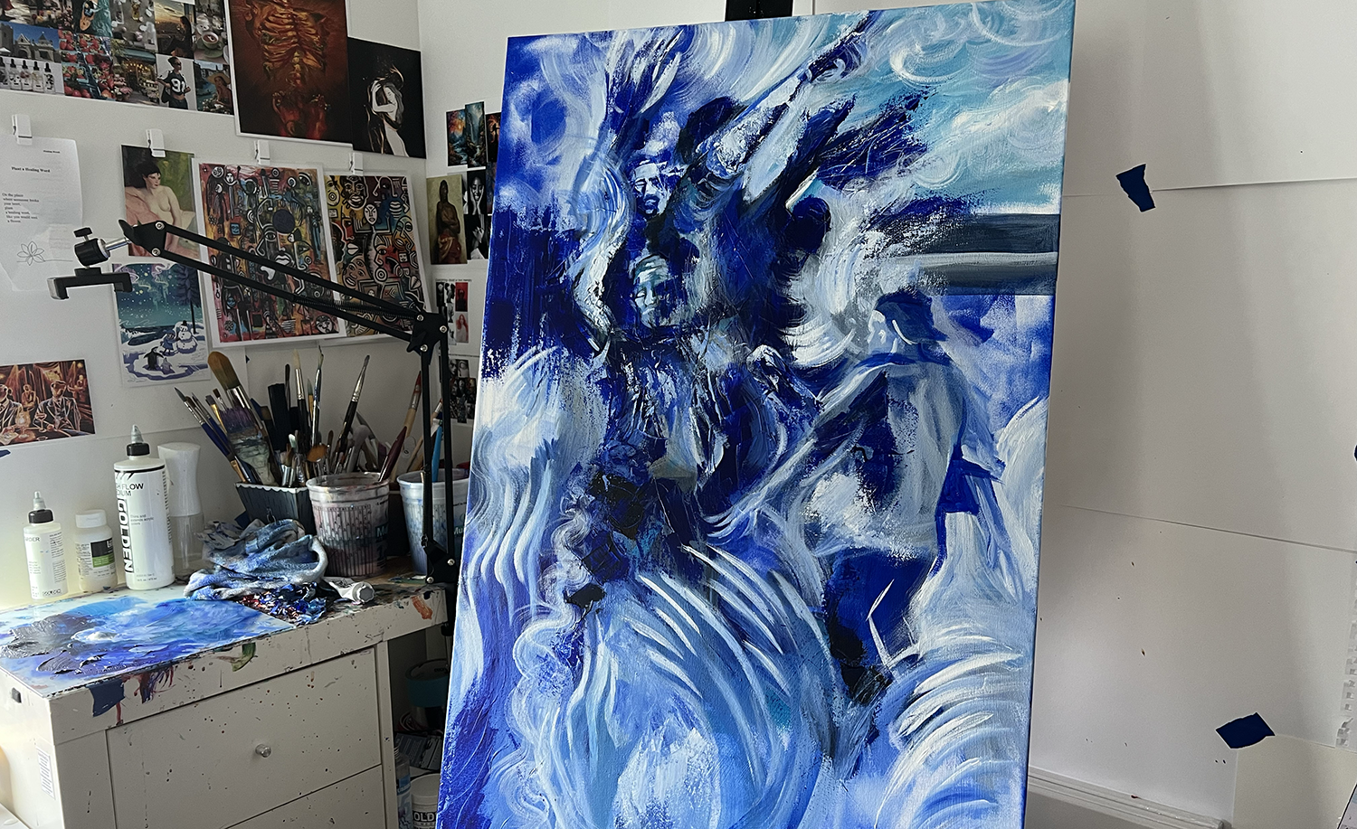 A monochromatic blue painting sits on the easel inside an artist's studio space. The studio has artwork on the walls, brushes on the table, and a messy glass palette on the table. This blue painting is a visual depiction of sound and musicians.