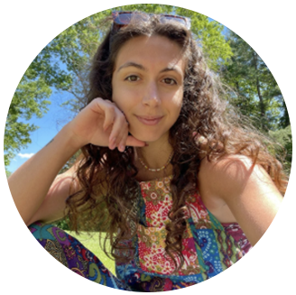 Photo of Morgan Foley- person of color with long curly brown hair and funky pattern on her romper sitting in the grass smiling