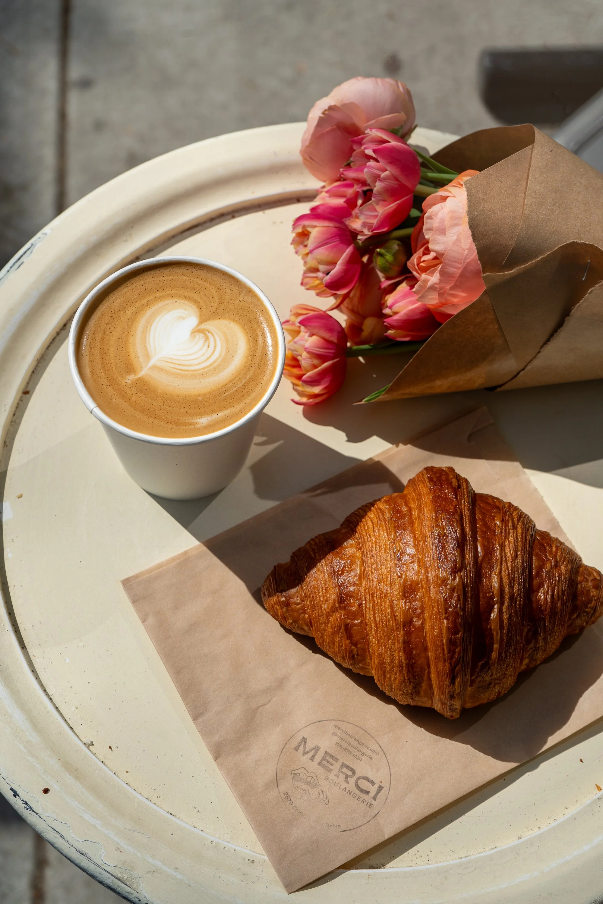 A cup of coffee with latte art, a croissant on a paper doily, a bouquet of pink flowers, and a paper bag of flowers on a white round table.