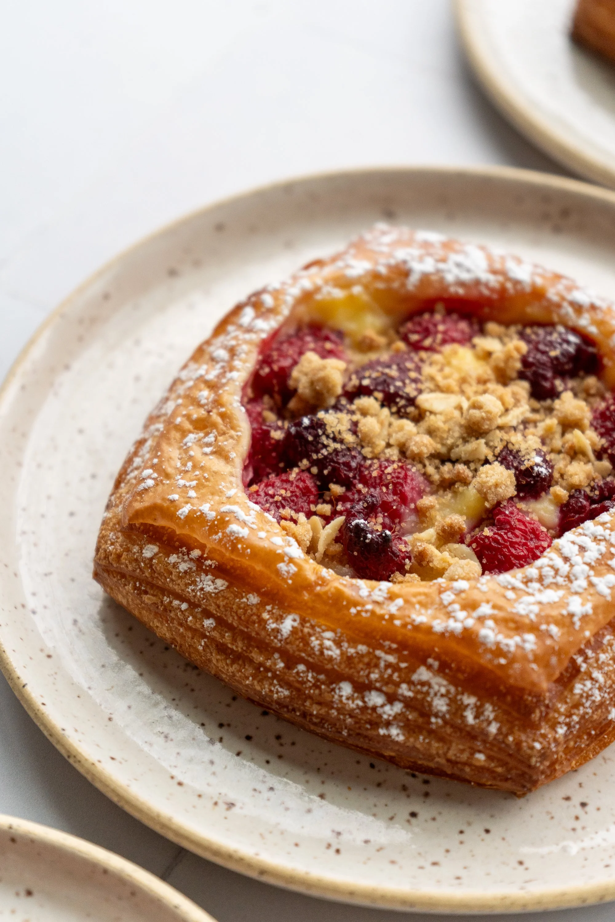 A close-up of a Danish pastry with mixed berries and crumb topping on a speckled beige plate.