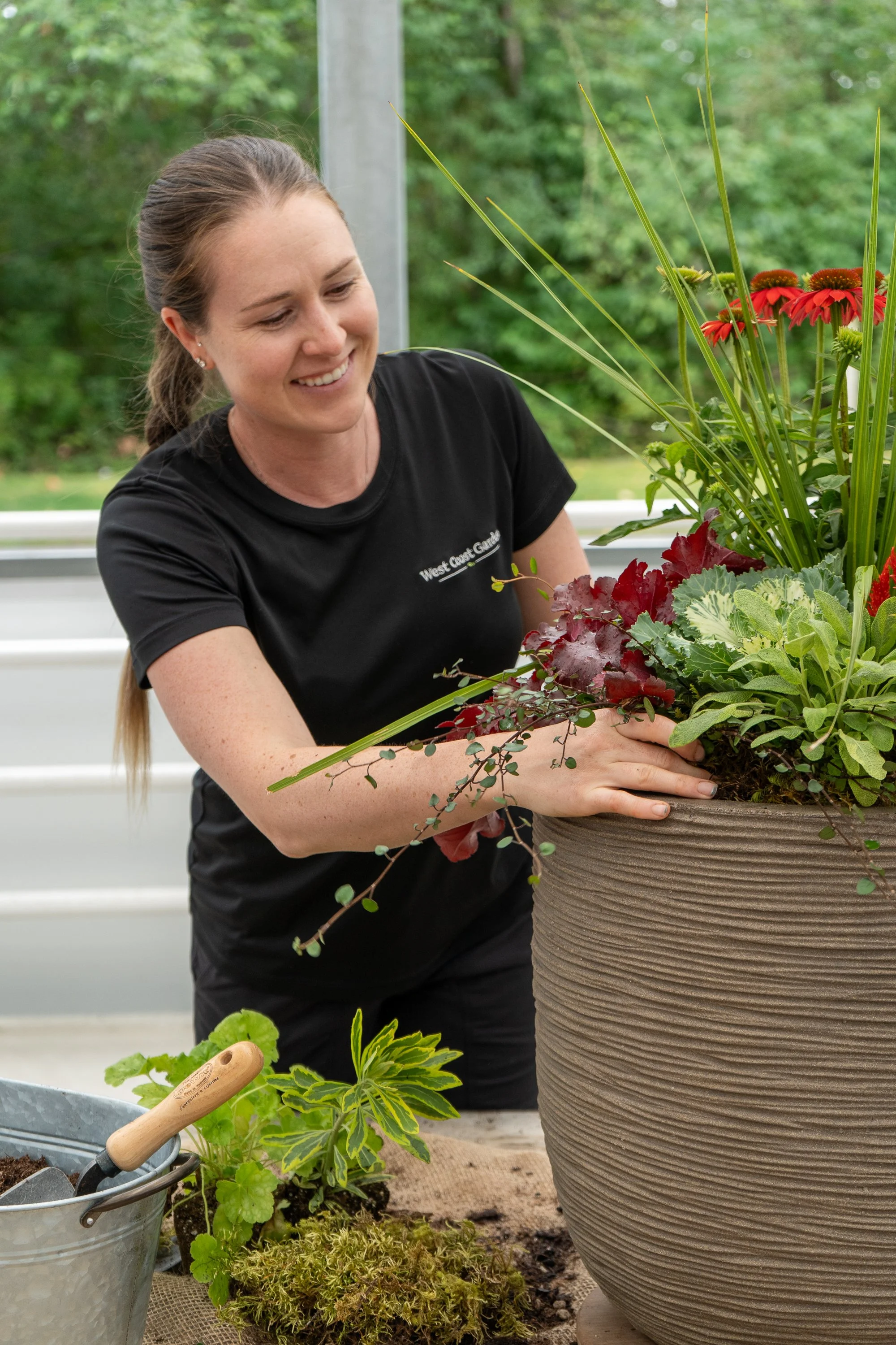 A woman is planting or arranging a large outdoor flower pot filled with various green and red plants and flowers, smiling as she works.