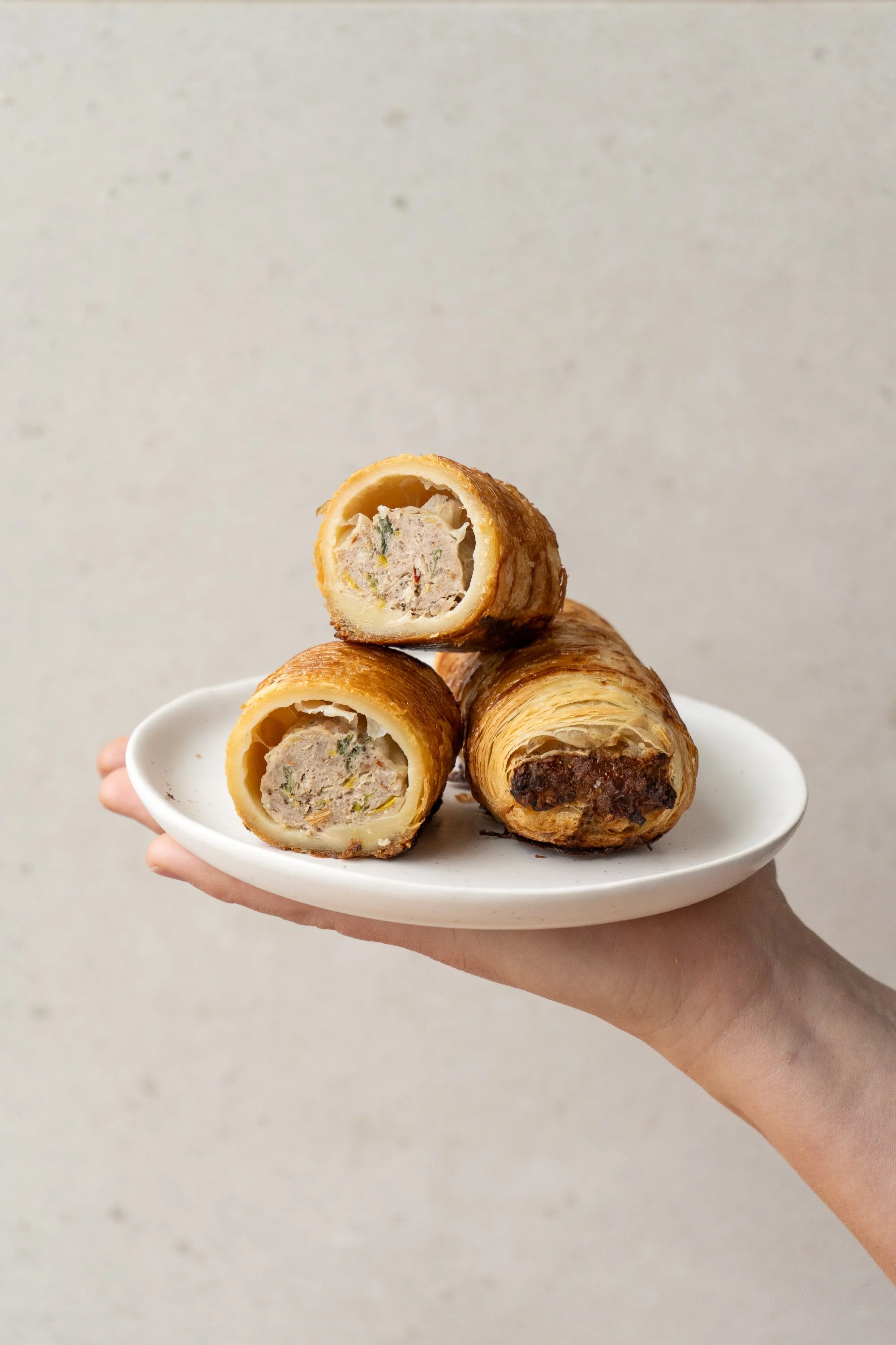 A hand holding a white plate with three savory pastry croquettes, two of which are cut open showing a creamy filling with pieces of vegetables and possibly meat, against a plain neutral background.