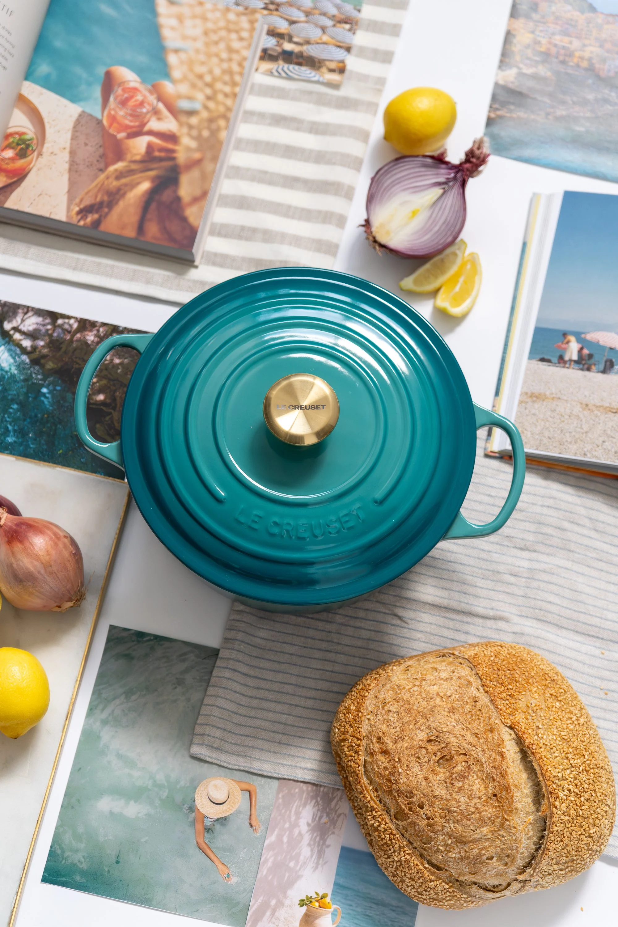 A teal Le Creuset cast iron pot with a gold knob on top, placed on a table among travel magazines, lemons, onions, and a loaf of bread.