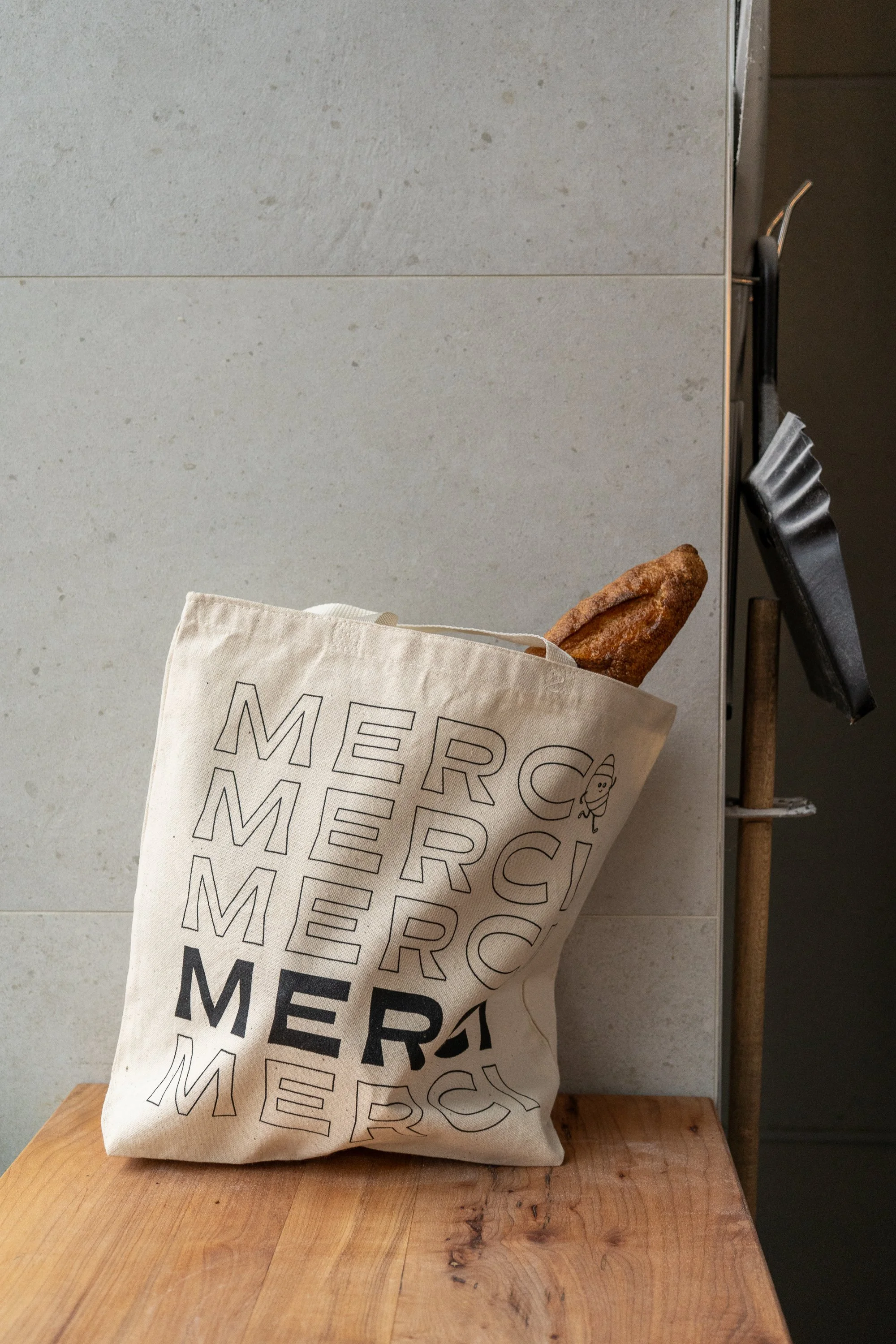 A beige tote bag with the word "MERCI" printed repeatedly in black, sitting on a wooden surface, with a breadstick peeking out of the bag, against a light gray tiled wall.